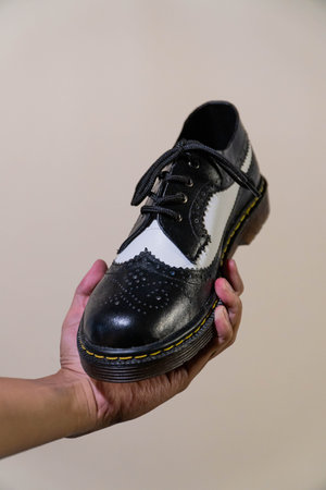A man's hand holds a black and white brogue wingtip shoe with a rubber outsole made from genuine cowhide. Men's hands holding elegant and shiny two tone shoes on a cream backgroundの写真素材