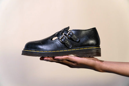 A man's hand holds black Mary Jane shoes with a rubber outsole made of genuine cow leather. Concept studio photo of hands holding elegant & shiny vintage shoes on beige backgroundの写真素材