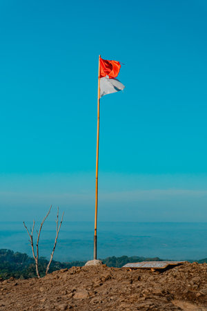 The peak of the ancient volcano Nglanggeran is dominated by rocks & contains the red and white Indonesian flag. Photo of rocky mountain peak with waving flag on a tropical morningの写真素材