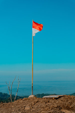 The peak of the ancient volcano Nglanggeran is dominated by rocks & contains the red and white Indonesian flag. Photo of rocky mountain peak with waving flag on a tropical morningの写真素材