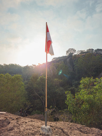 The peak of the ancient volcano Nglanggeran is dominated by rocks & contains the red and white Indonesian flag. Photo of rocky mountain peak with waving flag on a tropical morningの写真素材