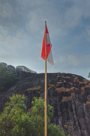 The peak of the ancient volcano Nglanggeran is dominated by rocks & contains the red and white Indonesian flag. Photo of rocky mountain peak with waving flag on a tropical morningの写真素材