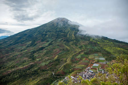 Mount Sindoro from a close distance with clouds around the peak on a sunny morning. The beautiful view of Mount Sindoro from the top of the hill at an altitude of 1800 mdplの写真素材