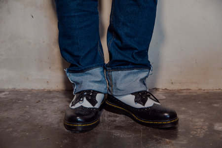 Close up of a man's legs wearing blue jeans paired with black & white brogue shoes. Close up of shiny two-tone wingtip brogue shoes against a dirty white wall.の写真素材