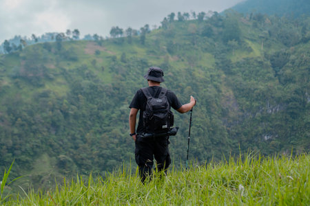 An adventurer posing with green hills background. Portrait of an Asian man with bucket hat, backpack, trekking pole and all black clothes hiking through beautiful green hills.の写真素材