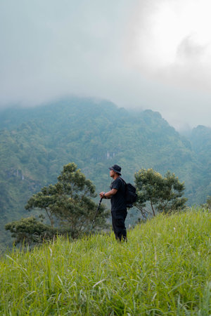 An adventurer posing with green hills background. Portrait of an Asian man with bucket hat, backpack, trekking pole and all black clothes hiking through beautiful green hills.の写真素材