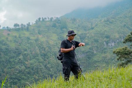 An adventurer posing with green hills background. Portrait of an Asian man with bucket hat, backpack, trekking pole and all black clothes hiking through beautiful green hills.の写真素材