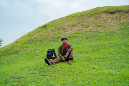 An adventurer is resting by sitting on a stretch of green grass hill. Asian male travelers enjoy the tranquility of nature by relaxing and enjoying the beautiful natural atmosphereの写真素材