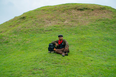 An adventurer is resting by sitting on a stretch of green grass hill. Asian male travelers enjoy the tranquility of nature by relaxing and enjoying the beautiful natural atmosphereの写真素材