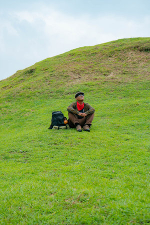 An adventurer is resting by sitting on a stretch of green grass hill. Asian male travelers enjoy the tranquility of nature by relaxing and enjoying the beautiful natural atmosphereの写真素材