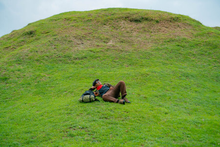 An adventurer is resting by lying down on a stretch of green grass. The Asian man enjoys the...
