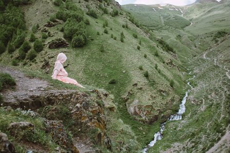 Little girl in pink hoodie sitting on the cliff and looking at the mountainsの写真素材