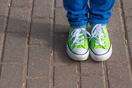 light green youth sneakers shod on legs in blue jeans, shot against the background of paving city tilesの写真素材