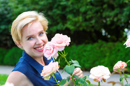 Blue-eyed blonde woman in a blue dress in the garden on a summer day admires delicate pink rosesの写真素材