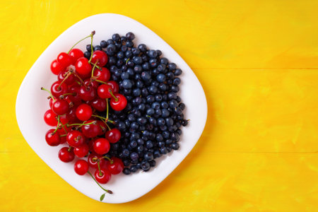 berries of cherries and blueberries on a plate, summer seasonal berries, on a yellow wooden backgroundの写真素材
