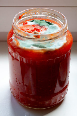 A jar in which bright red tomato sauce is covered with a thick layer of green and white mold stands...