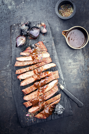 Traditional American barbecue dry aged flank steak sliced with hot sauce and chili as top view on an old carbonized boardの写真素材