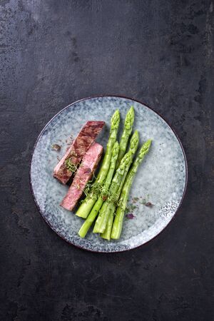 Barbecue dry aged wagyu roast beef steak with blanched green asparagus and herbs as top view on a modern design plate with copy spaceの写真素材