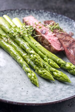 Barbecue dry aged wagyu roast beef steak with blanched green asparagus and herbs as closeup on a modern design plate with copy spaceの写真素材