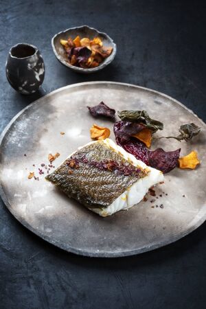 Gourmet fried European skrei cod fish filet with algae and vegetable crisps as closeup on a modern design plateの写真素材