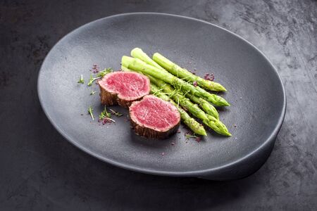 Barbecue dry aged wagyu beef fillet head medallion steak natural with green asparagus and herbs as closeup on a modern design plate with copy spaceの写真素材