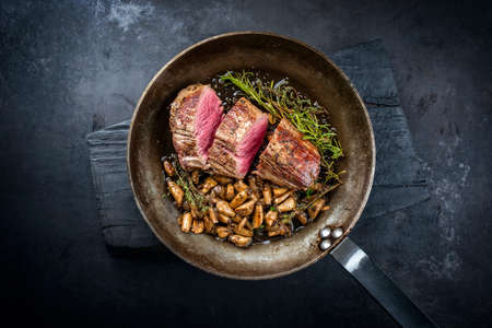 Fried dry aged beef fillet steak natural with king trumpet mushroom and herbs offered as top view in a rustic frying panの写真素材