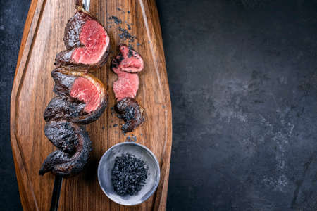 Modern style barbecue dry aged wagyu Brazilian picanha from the sirloin cap of rump beef sliced and offered as top view on a skewer on a wooden design board on rustic black background with copy space rightの写真素材