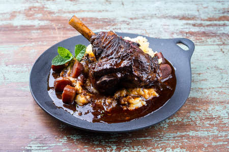 Modern style traditional braised slow cooked lamb shank in red wine sauce with shallots and mashed potatoes offered as close-up in a design cast iron plateの写真素材