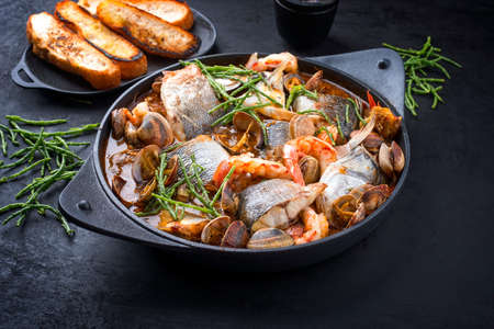 Modern style traditional Spanish seafood zarzuela de pescado with fish, king prawns and venus clams served with barbecue garlic bread in red sauce as close-up in design potの写真素材