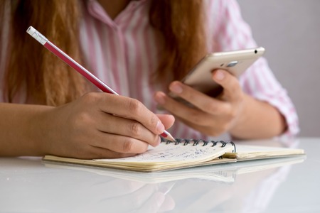 Girl With Mobile Phone Writing On Paper At Table In Classroom. Vertical view of a student texting during exam.の写真素材