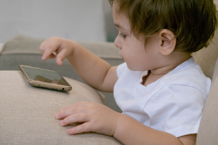 Adorable toddler boy sitting on the sofa in the living room and playing with smartphone. Child learning how to use smartphone. Boy texting on the phone. - technology and lifestyle conceptの写真素材