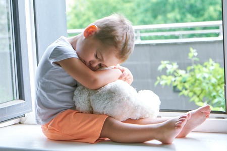 boy of two years sitting by the window and hugs a toy Bunny. rainy weather, waiting for dad to come home from workの写真素材