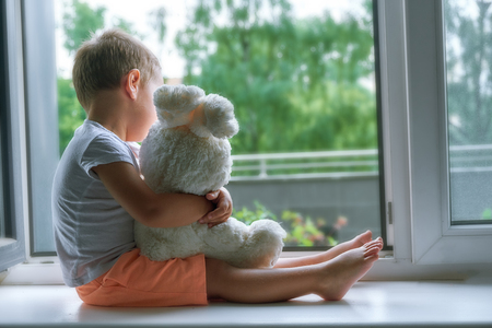 boy of two years sitting by the window and hugs a toy Bunny. rainy weather, waiting for dad to come home from workの写真素材