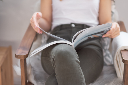Beautiful girl at home sitting on the couch, reading a magazine and having a coffee break, relaxation conceptの写真素材
