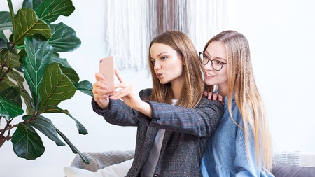 Happy young two cheerful women sitting in the sofa taking a selfie on the mobile phoneの写真素材