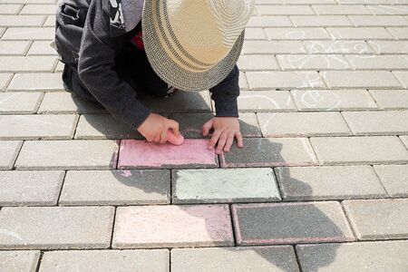 child drawing with chalk on asphalt. boy playing alone in the street. holding a piece of chalk in his fingersの写真素材