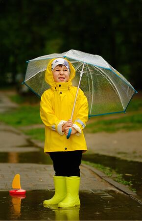 Cute child playing in a puddle with a ship on a summer rainy day. a boy in a yellow raincoat walks in the park.の写真素材
