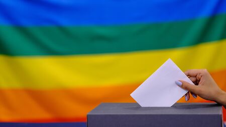 Hand holding ballot paper for election vote concept. elections, The hand of woman putting her vote in the ballot box. Rainbow flag on backgroundの写真素材