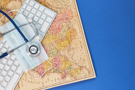 Keyboard, medical stethoscope and a protective mask on a geographical map. Concept stay home and...