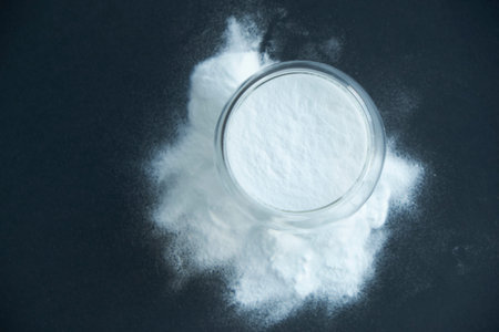 Glass bowl of baking soda. Spoonful of bicarbonate. Top view, flat lay. Close up on a black...