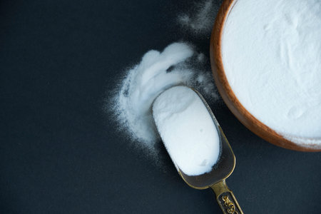 Glass bowl of baking soda. Spoonful of bicarbonate. Top view, flat lay. Close up on a black...