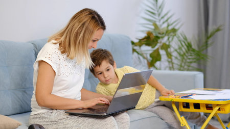 Smiling mom working at home with her child on the sofa while writing an email. Young woman working...