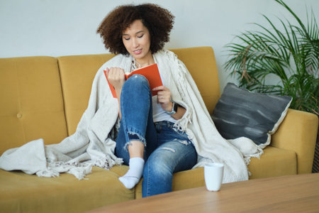 Beautiful young African American woman reading a book lying comfortably on her back on a couch in...