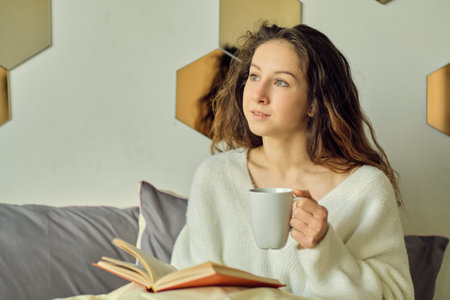 woman sits in bed and drinks coffee. enjoying the early morning with a book in hand. cozy winter...