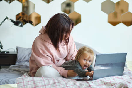 mom working remotely on laptop while taking care of her baby. Young mother on maternity leave trying to freelance by the desk with toddler child. Close up, copy space, background.の写真素材