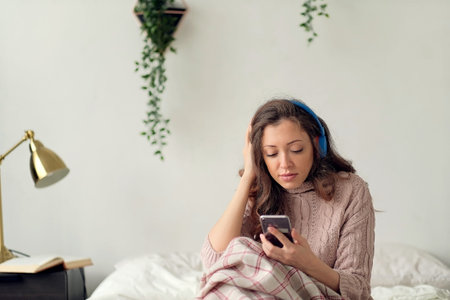 Young cheerful woman lying on bed shuffling songs on mobile phone. Relaxed girl listening music with...