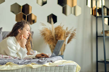 Smiling young woman wearing headphones lying on bed with laptop, teenage student studying online at home, listening to audio course, fast learning foreign language at home, head shot portraitの写真素材