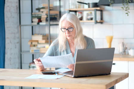 Senior woman using laptop for websurfing in her kitchen. The concept of senior employment, social security. Mature lady sitting at work typing a notebook computer in an home office.の写真素材