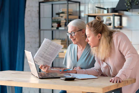 Senior woman using laptop for websurfing in her kitchen. middle-aged daughter helps her mother with documents. Mature lady sitting at work typing a notebook computer in an home office.の写真素材