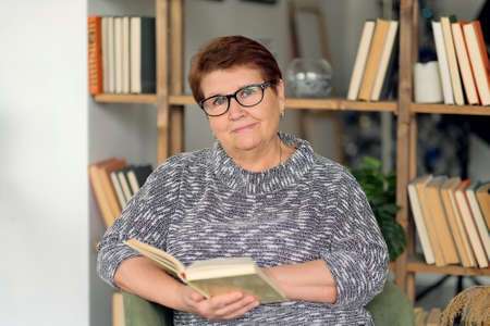 Beautiful old woman in casual clothes reading on couch at home. Beautiful old woman in casual clothes reading on couch at home. Senior woman with poor eyesight in glasses peers into a book.の写真素材